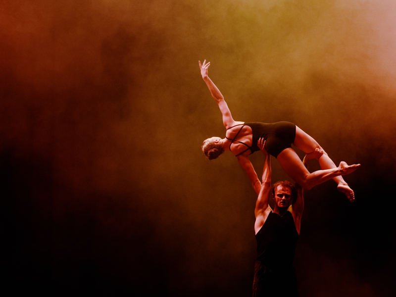 A male dancer lifts a female dancer above his head, in front of a dark and moody amber smoke filled background