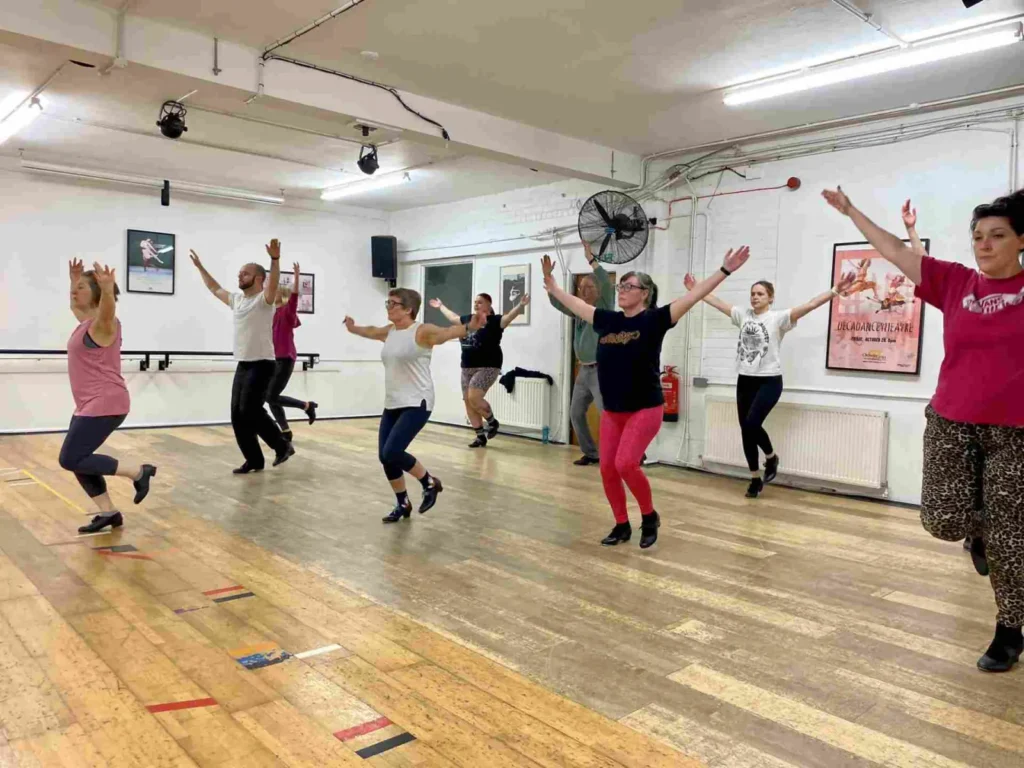 Adult beginners in a tap class at Hype Dance's Sheffield studio, with their arms raised and one foot lifted mid-step